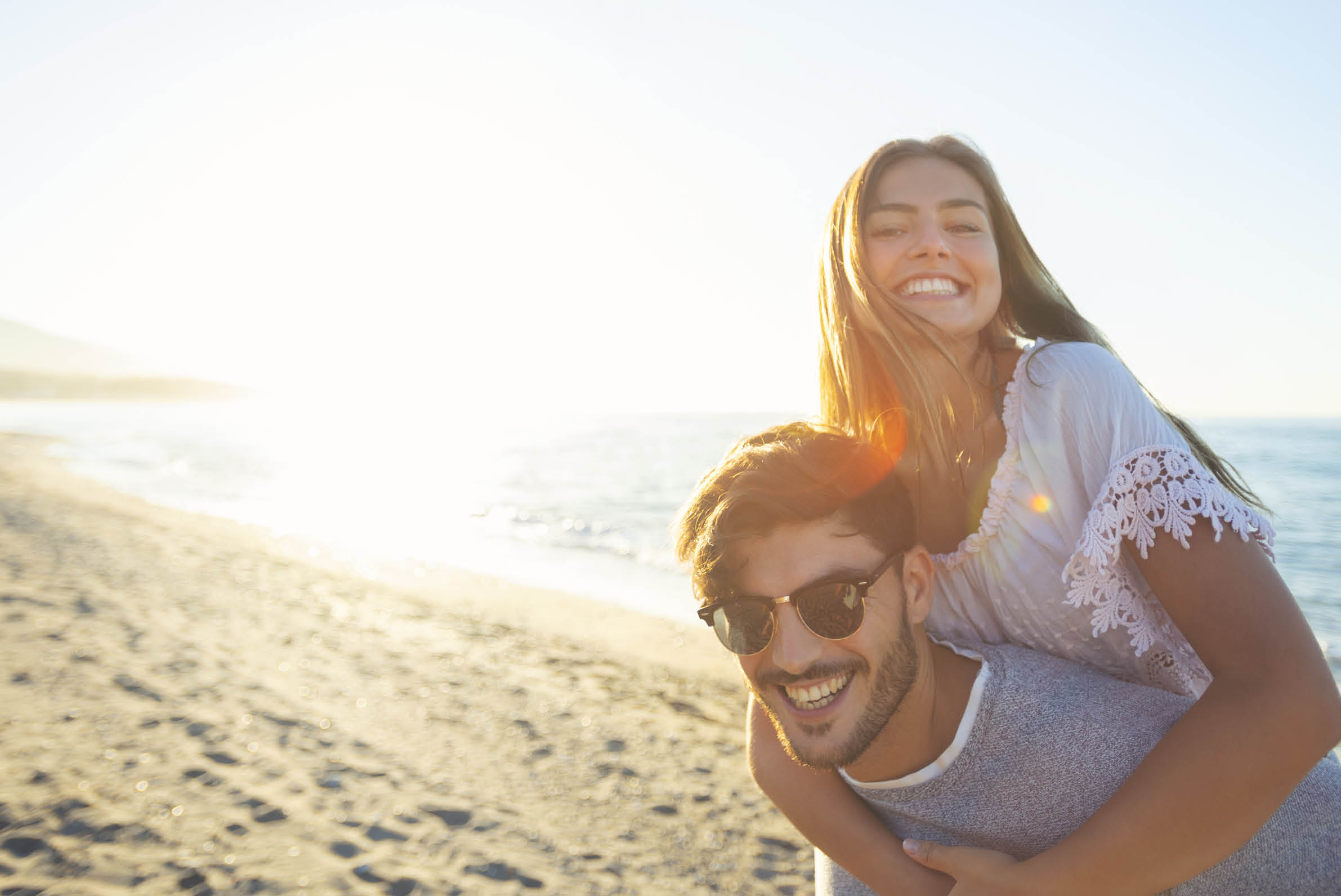 Couple playing piggyback on the beach at sunset or sunrise. They are happy, smiling and laughing having fun with the ocean in the background.