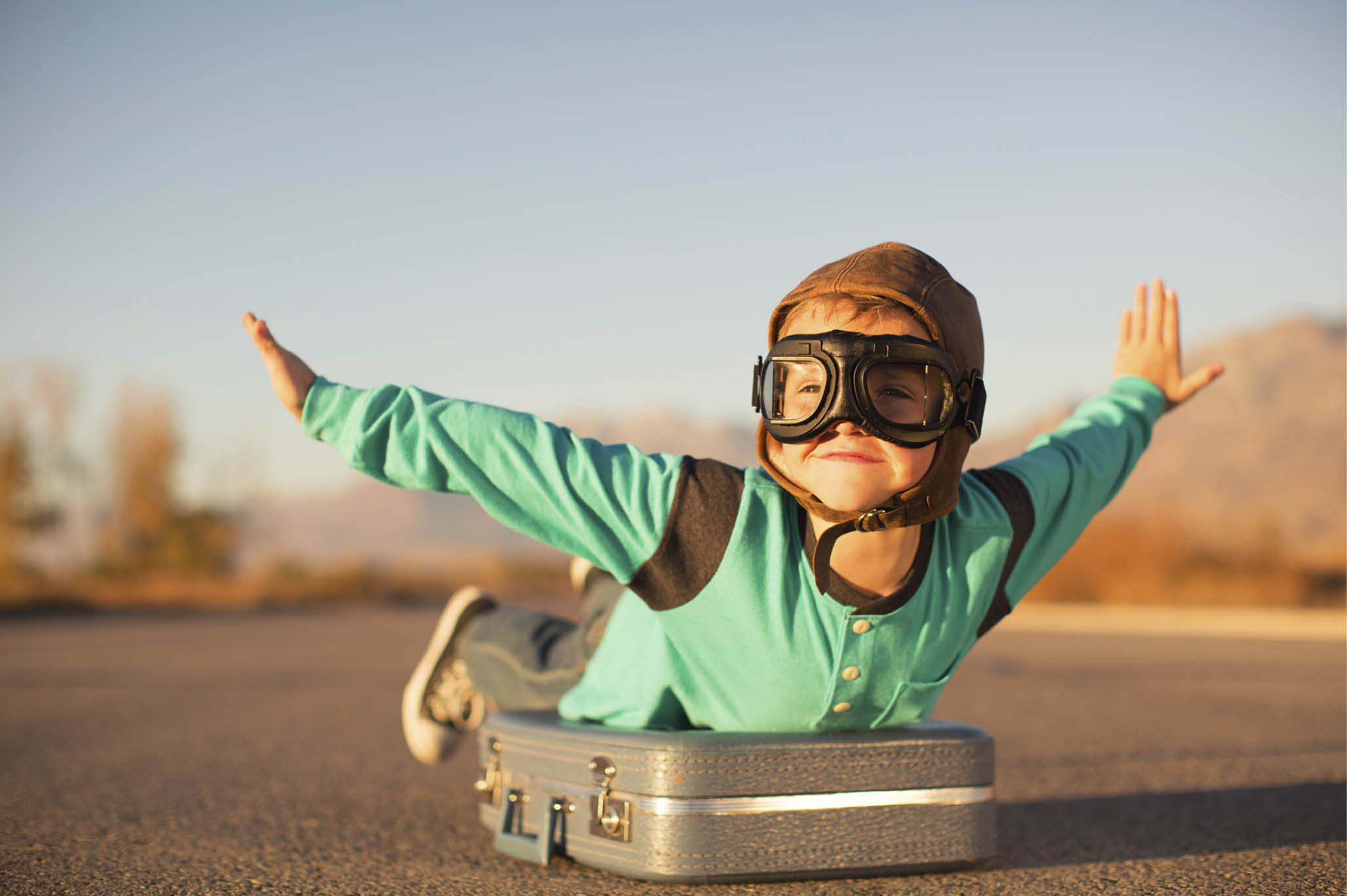 A young boy with outstretched arms lies on top of a suitcase imagining he is flying on an airplane away and traveling to exotic locations. He is wearing a flight cap and goggles and has a large smile on his face. He loves to travel. 